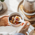 A person holds a small white bowl filled with a warm herbal soup or dessert, using a spoon to lift a piece. A glass beverage maker sits in the softly blurred background on a wooden tray, with a plant and neutral fabric visible around it.
