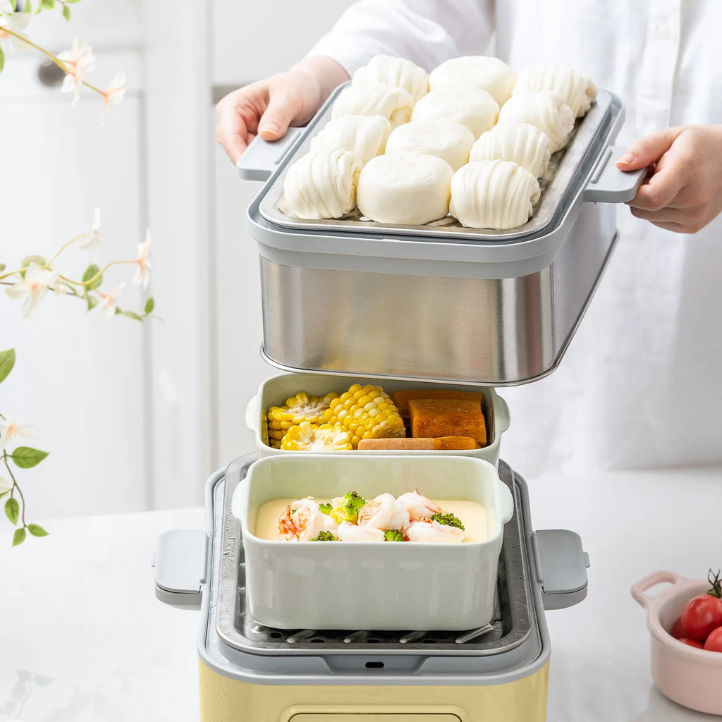 A person in a white shirt is holding a steamer insert loaded with white steamed buns above a multi-compartment steamer basket containing corn and shrimp dumplings, set on a light countertop.