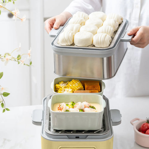 A person in a white shirt is holding a steamer insert loaded with white steamed buns above a multi-compartment steamer basket containing corn and shrimp dumplings, set on a light countertop.